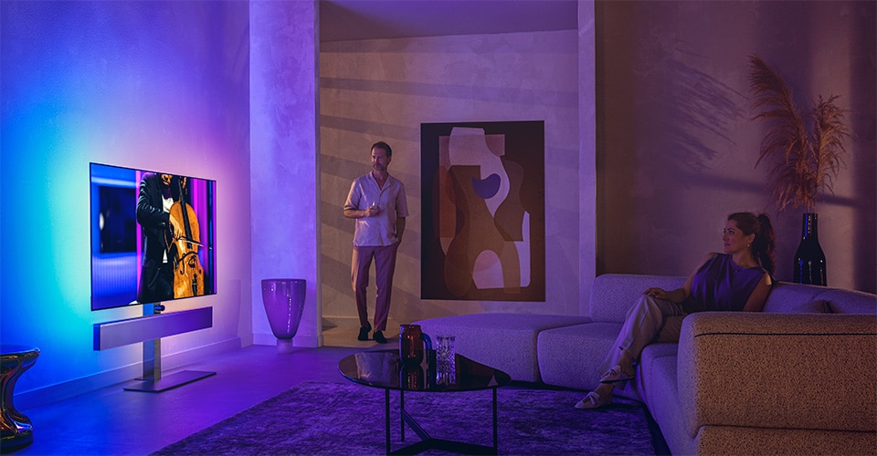 Homem e mulher numa sala de estar elegante assistindo a uma TV Philips Ambilight exibindo uma apresentação de violoncelo.