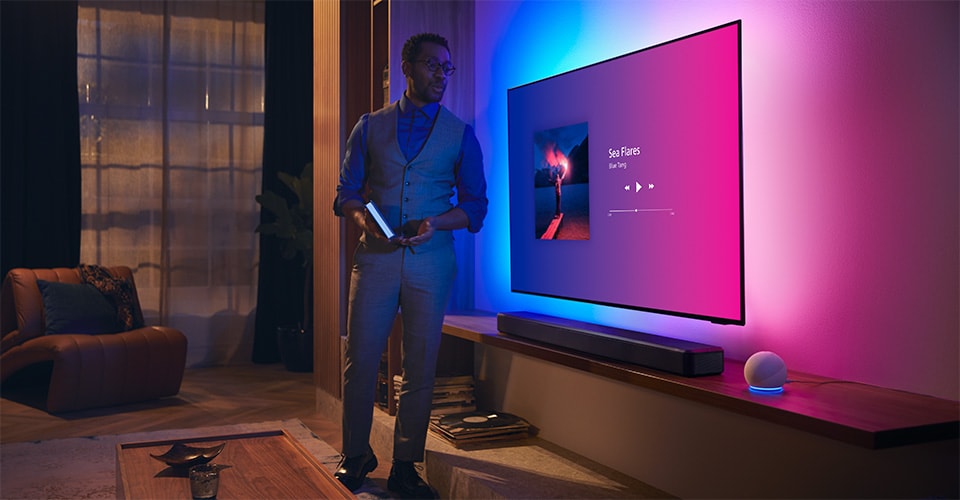 Pessoa numa sala de estar moderna perto de uma TV Philips Ambilight com interface de música e barra de som Fidelio