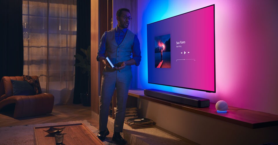 Pessoa numa sala de estar moderna perto de uma TV Philips Ambilight com interface de música e barra de som Fidelio