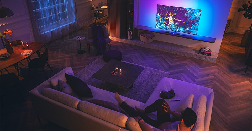 Homem relaxando num sofá numa sala de estar com pouca iluminação, assistindo a uma TV Philips Ambilight.