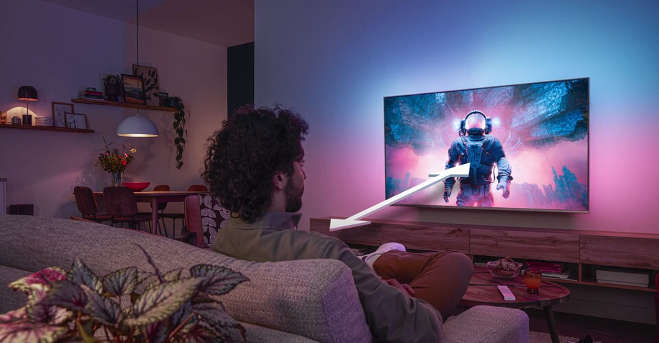 Homem sentado num sofá assistindo a uma TV Philips Ambilight exibindo uma cena colorida com um astronauta, com uma seta indicando a distância ideal de visualização.