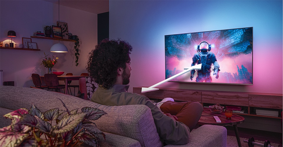 Homem sentado num sofá assistindo a uma TV Philips Ambilight exibindo uma cena colorida com um astronauta, com uma seta indicando a distância ideal de visualização.