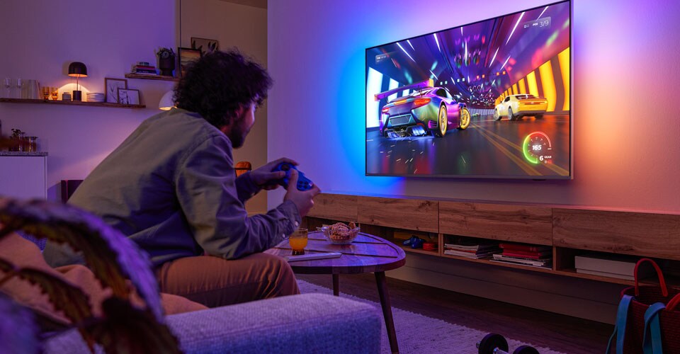 Uma pessoa está sentada no sofá, a jogar um videojogo de corridas numa TV Philips Ambilight.