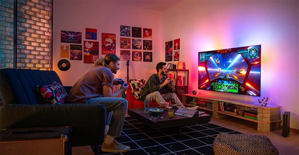 Duas pessoas jogam com entusiasmo um videojogo com tema espacial numa sala de estar acolhedora e colorida.